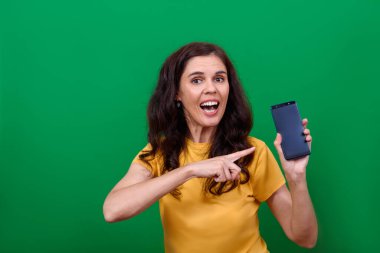 Genç ve çekici bir kadın elindeki boş telefon ekranını işaret ediyor ve kameraya bakıyor.