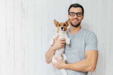 Çekici neşeli Kafkas erkek casual t shirt, gözlükler, favori Evcil hayvan tutar, Beyaz ahşap arka plan boşluk ile karşı duruyor. Onun mutlu tıraşsız adamla jack russell terrier