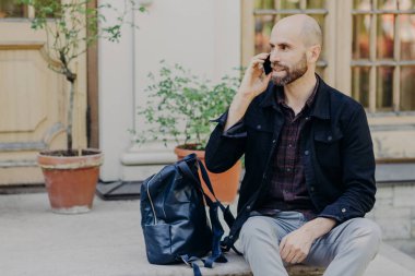 Yakışıklı kel erkek sakallı, uzun yürüyüş sonra açık kalan vardır, cep telefonu bir yana, odaklı, sırt çantası ona pozlar builduing arka plan karşı yakın biliyor. İnsanlar ve iletişim kavramı