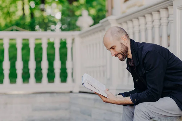 Photo of middle aged bald bearded man leans closely to book, has positive expression, sits against white balcony, has dark stubble, being in good mood as likes reading. Male reads romantic story