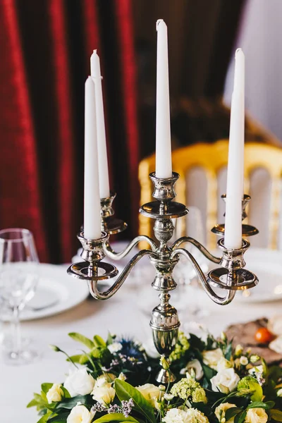 Luxurious banquet table served with beautiful candlestick with white candles, nice flowers, tablecloth and dishes. Retro style restaurant. Wonderful elements of decor. Table prepared for celebrating