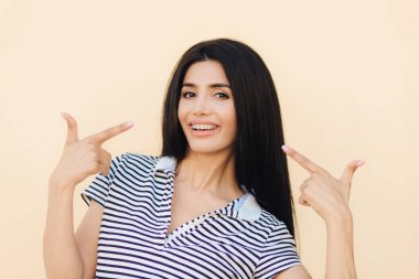 Memnun gülümseyen esmer kadın ağzında gösterir, pozitif ifadesi olan, gösterir ağız geniş gülümsemeyle, uzun siyah saçlı, çizgili t gömlek giymiş pembe arka plan üzerinde ıssız