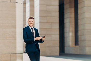 Pozitif başarılı erkek girişimci lüks siyah takım elbiseli, modern tablet tutar, mutlu bir yere benziyor, modern ofis binası içinde duruyor, Internet veya sosyal ağlar iş molası sırasında arar