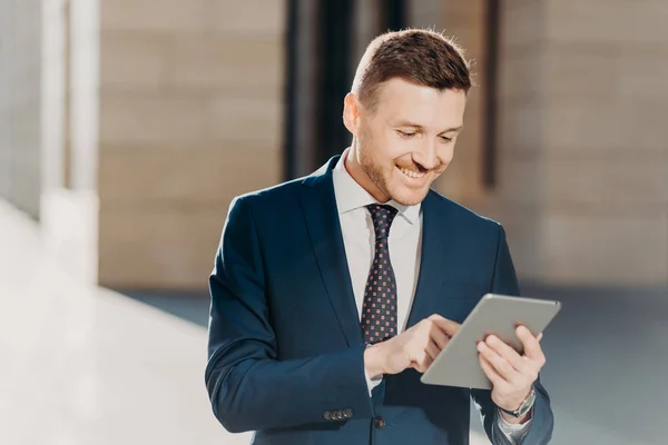 Happy prosperous businessman in formal wear, makes money transaction via touch pad, has glad expression, stands outdoor, connected to wireless intrnet. People, business and technology concept