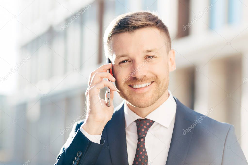 Portrait of delightful European businessman, wears elegant suit, talks on smart phone, deals with business partner, decide time of meeting, has positive expression. People, career, technology concept