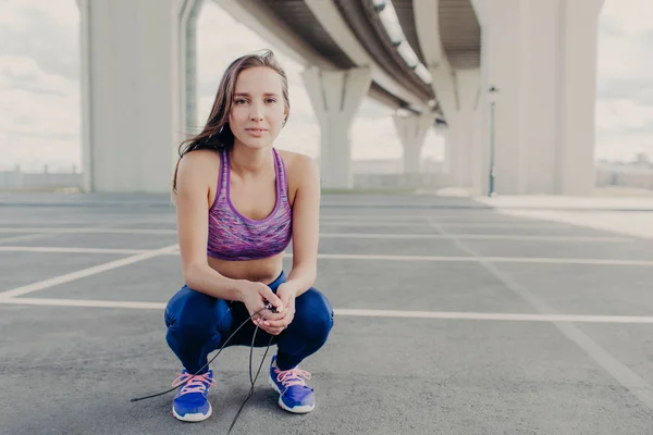 People, youth and sport concept. Self assured slim female goes in for sport, jumps with skipping rope, sits down, likes physical activities. Beautiful young fit woman in sport top and sneakers