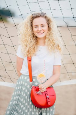 İyi görünümlü kıvırcık kadın dikey atış t gömlek ve etek giyer, küçük çanta, buzla kokteyl tutar, tenis net plaj yakınında standları, iyi dinlenme vardır. Çekici çekici gülümseme ile güzel kadın