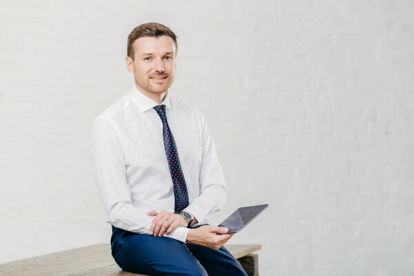 Pleased male entrepreneur makes payment online on digital tablet, dressed in elegant white shirt, tie and trousers, sits on wooden table, isolated over white background. Business concept