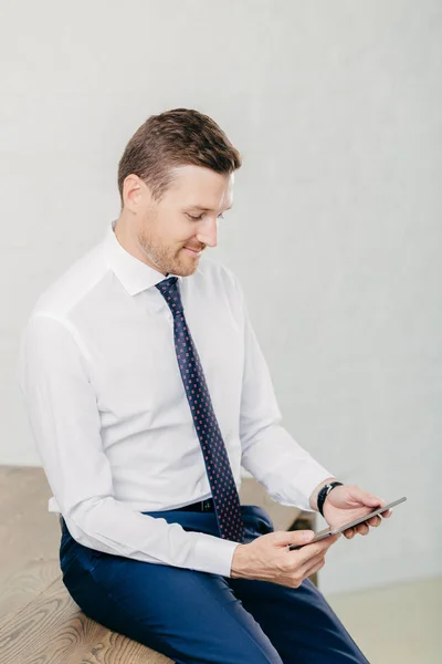 Young unshaven intelligent male wears formal clothes, uses digital tablet for work, focused into screen, sits at office desk, isolated over white background. Programmer develops application software