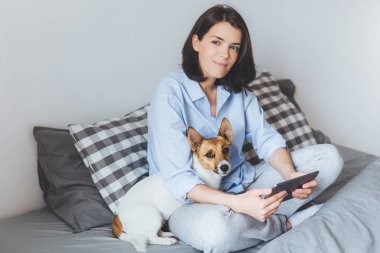 Rüya gibi esmer kadın modeli pijama çapraz bacaklar rahat yatak yatak odasında oturur, modern tablet tutar, onu evde beslenen hayvan kucaklayan, açık havada yürüyüş hakkında rüyalar. İnsanlar, dinlenme, teknoloji