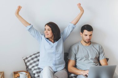 Genç evli çift sabah yatakta geçirmek: güzel esmer erkek uzanır ve kocası ciddi ifade ile dizüstü bilgisayar kayıtsızca, giyinmiş uzaktan çalışma için yatak odasında olmak kullanır