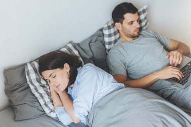Meşgul erkek yatakta yatıyor, dizüstü bilgisayarda çalışır, karısı sağlıklı uyku, yatak odasında birlikte olmak varken ciddi ifade etti. Yatakta iki kişi. İnsanlar, Aile, ev concept