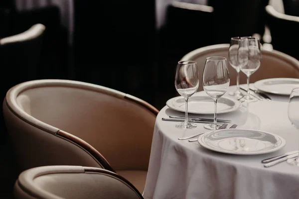 Cutlery and wine glasses on white tablecloth, empty armchairs near. Banquet hall in restaurant. Served table for guests in cafe. Celebration concept