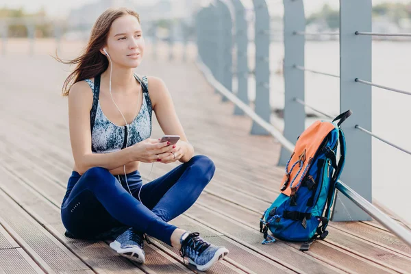 Dreamy attractive female wears top and leggings, thinks about something while listens music, holds modern cell phone, bag stands near, breathes fresh air in morning. Recreation and lifestyle concept