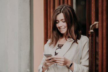 Koyu saçlı, beyaz zarif ceketli giyinmiş genç gülümseyen kadın modern akıllı telefon tutar, Ofis Binası, sosyal ağlarda arkadaşlarınızla iletileri kapılardan yakın duruyor. Teknoloji kavramı