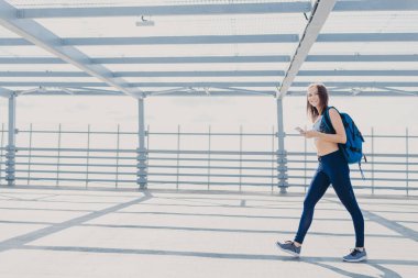 Aktif kadın fitness eğitmeni sportsclothes, spor ayakkabı giymiş, sırt çantası taşıyan, kalori kontrol etmek için cep telefonu kullanır, kulaklık ile müzik dinler, ücretsiz Wi-Fi bağlantı kullanır. İnsanlar ve spor