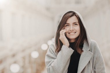 Gençlik ve modern teknolojileri kavramı. Memnun esmer Avrupa kız sıcak hoodie giyer, cep telefonu üzerinden iletişim sahiptir, sevinçle mesafe içine görünüyor, hoş bir şey adam ile tartışır