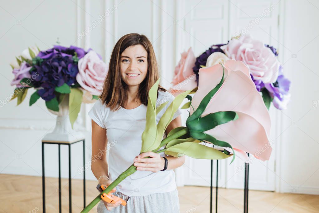 Floral design concept. Beautiful satisfied female florist makes composition of beautiful artificial flowers, decorates hall, has positive smile, dressed casually, being photographed at work.