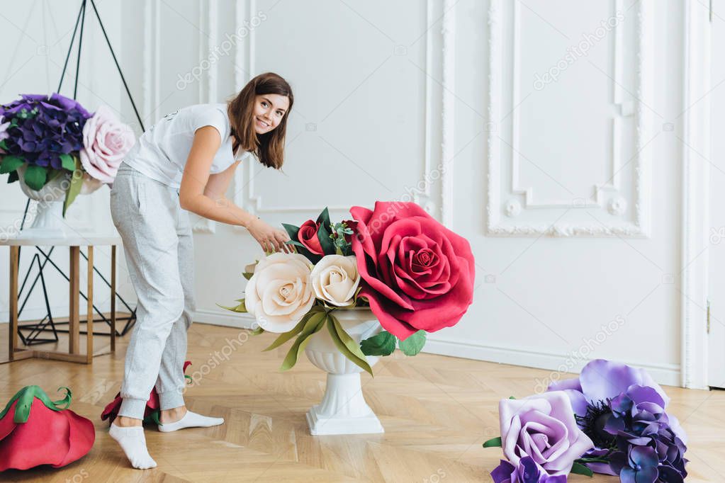 Busy woman dressed casually, decorates hall with flowers for festive event. Female florist prepares banquet hall to wedding, uses different flower compositions