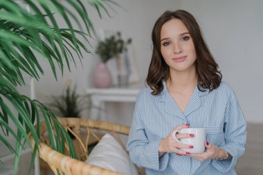 Photo of pretty female with appealing look, dressed in casual domestic clothes, holds white mug with aromatic coffee or tea, sits on wicker furniture indoor with copy space for your advertisement
