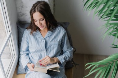 Üstten görünüm kitap haline duruldu karanlık uzun saçlı memnun kadın heyecan verici bir şey okur, rahat bir atmosfer evde sahiptir, pencere pervazına boş odada teşkil etmektedir. İçin zaman okuma ve dinlenme.