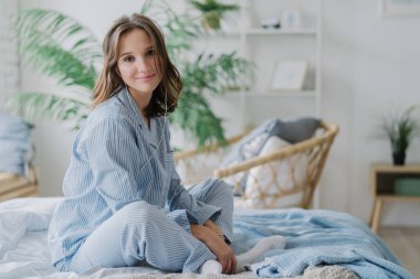 Oldukça koyu saçlı kadın kapalı atış çapraz bacakları yatak, nighclothes, beyaz çorap giymiş oturur, direcltly kameraya benziyor, dinlediği müzik kulaklık içinde sakin iken rahat hissediyor. Dinlenme ve izin günü