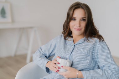 Headshot ile beyaz bir kadın, makyaj, pijama giymiş, çay bir fincan tutar, rahat koltuğa oturur, iyi dinlenme evde sahiptir, kamera çekici ifade ile görünüyor. Aile içi atmosfer