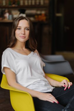 Heathy deri, koyu saçlı güzel kadın hoş görünüm gündelik kıyafeti giymiş, koltukta, rahat hissediyor, karanlık odada teşkil etmektedir. Memnun kız kamera memnun ifade ile poz veriyor