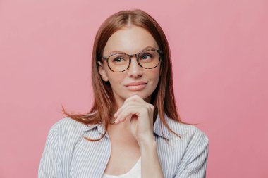 Düşünceli iş kadını çene tutar, bir kenara dalgın ifade ile görünür, gözlük takıyor, gömlek giymiş siyah saçlı, yeni strateji geliştirilmesi üzerinde düşünüyor, pembe studio arka plan üzerinde modelleri