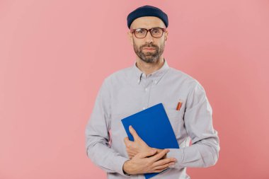 Shot ciddi tıraşsız genç Avrupa erkek güvenle kameraya benziyor, karanlık kıl vardır, kayıt, resmi gömlek ve şapkalar, pembe bir arka plan karşı modelleri giyinmiş belirterek için Not defteri tutar.