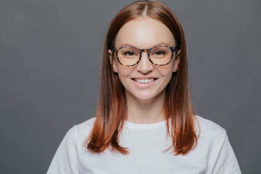 Headshot hoş görünümlü koyu saçlı Genç kadının içinde rahat kıyafetler giymiş şeffaf gözlük takar, beyaz mükemmel dişler, gri arka plan üzerinde izole gösterir, sağlıklı cilt, olumlu bir görünüm vardır.