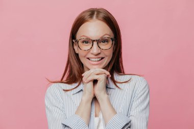 Pozitif Genç beyaz bir kadın eller çene altında tutar, siyah saçlı, dişlek gülümseme, beyaz gömlek ve gözlük takıyor, pembe bir arka plan üzerinde modeller, bir şey büyük ilgi ve coşku ile dinler