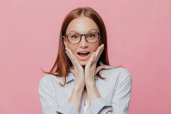 Human facial expressions, reaction concept. Beautiful prosperous business lady keeps both hands on cheeks, wears elegant white striped shirt, has surprised facial expression, isolated over pink wall