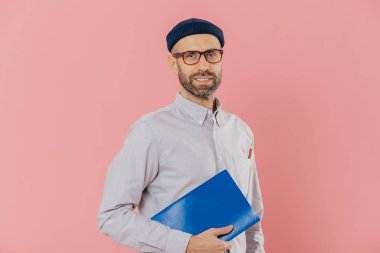 İnsanlar ve eğitim kavramı. Sakallı Genç beyaz adam öğrenci ders kitabı tutan, son sınav için hazırlar, Gözlük, şapka ve resmi gömlek, pembe bir arka plan karşı yan duruyor giyer