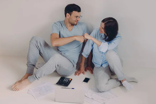 Positive female and male coworkers give fist bump to each other, work as team, calculate figures and prepare financial report, pose on floor, isolated over white background. Couple plan family budget