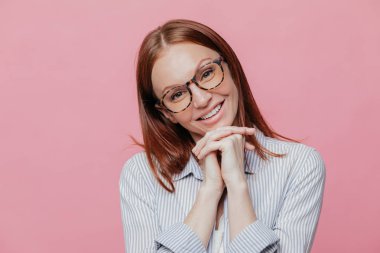 Zarif güzel kadın kafa tilts, eller çene altında tutar, şeffaf gözlük ve gömlek, gülümsüyor yavaşça giyer, pembe bir arka plan üzerinde modeller. İnsanlar ve olumlu duygular kavramı. Büyüleyici kadın