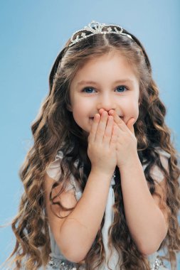 Görüntü olumlu güzel gülümseyen çocuğun dudaklarından iki elini tutar, uzun kalın saç, mavi gözleri var, taç giyer, mavi arka planı poz veriyor, mutluluk ifade eder. Çocuk ve memnuniyeti