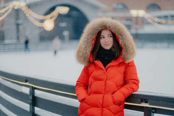 Foto van mooie doordachte jonge vrouw draagt rode winterjas met bont op hoody, houdt de handen in de zakken, staat tegen onscherpe achtergrond in de buurt van ijsbaan, heeft buiten lopen in de winter — Stockfoto