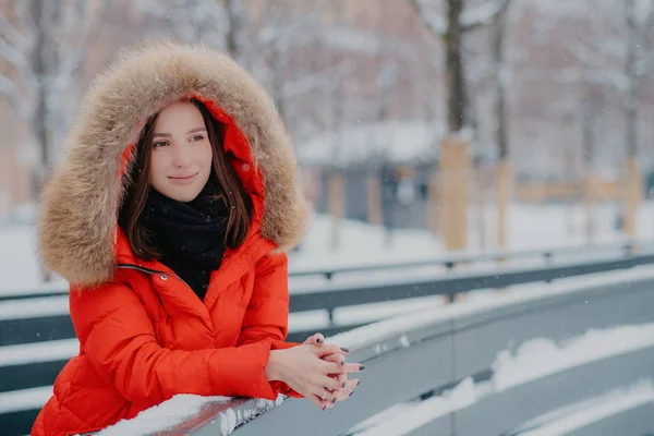 Outdoor shot van doordachte Europese vrouw draagt rode winter jas met hoody op hoofd, leunt op vandaar pensively onderzoekt op afstand, geniet goede frosty besneeuwde weer, manicure heeft. Levensstijl — Stockfoto