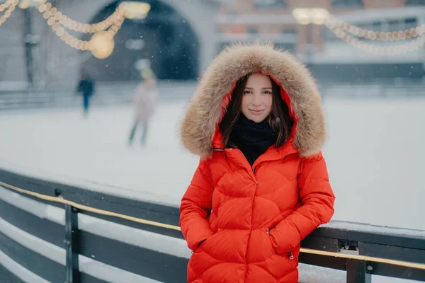 Aantrekkelijke jonge vrouw in rode warme jas, staat in de buurt van ijsbaan, gonna skate, houdt de handen in de zakken, kijkt met zachte glimlach direct camera, besteedt weekend actief. Winter tijd concept — Stockfoto