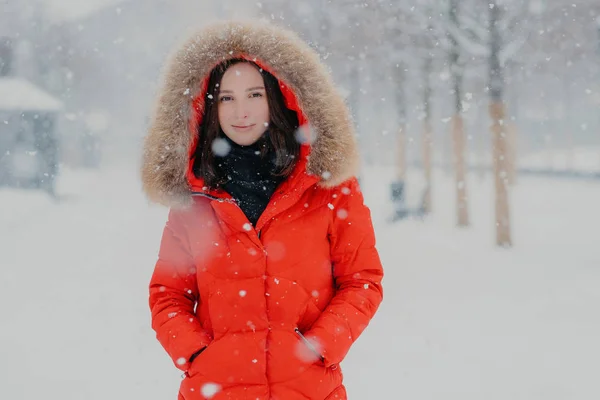 Mooie vrouwelijke model in rode jasje, stads buiten tijdens de sneeuwval, camera, gonna hebben wandeling met vriendje met donkere ogen kijkt, ademt van frisse lucht. Mensen, wintertijd en vrije tijd concept — Stockfoto