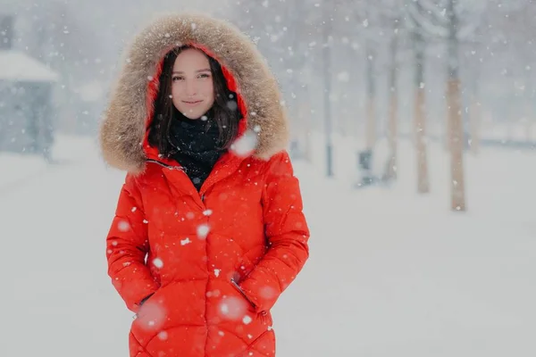 Buiten schot van aantrekkelijke vrouw gekleed in winterkleren, houdt beide handen in de zakken, kijkt met tevreden expressie direct bij de camera, wandelingen in de open lucht tijdens de sneeuwval. Besneeuwde weer. — Stockfoto