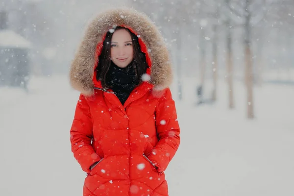 Goed uitziende jonge vrouw heeft aantrekkelijke look, warme rode jas draagt, houdt de handen in de zakken, heeft buiten wandeling tijdens wintertijd en sneeuwval, vormt op de achtergrond wazig sneeuw. Seizoen concept — Stockfoto