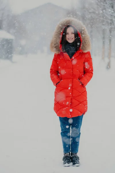 De verticale shot van de volledige lengte van mooie vrouw draagt rode winter jas, jeans en laarzen, houdt de handen in de zakken, staat op straat tijdens wintertijd sneeuwvlok, vormt voor het maken van foto buiten — Stockfoto
