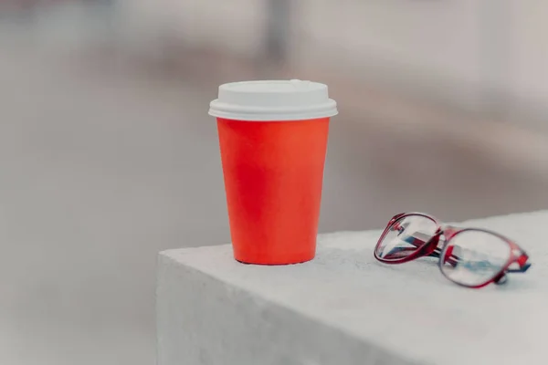 Horizontal shot of red coffee to go and optical glasses. Aromatic coffee for you to drink. Drinking concept. Paper cup of hot beverage
