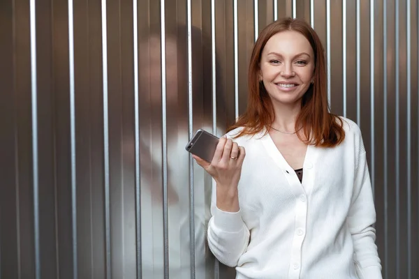 Симпатичная деловая женщина ждет звонка, держит современный мобильный телефон, одетый в белый свитер, имеет каштановые волосы, подключен к беспроводному интернету, пользуется передовыми технологиями, получает уведомление от друга — стоковое фото
