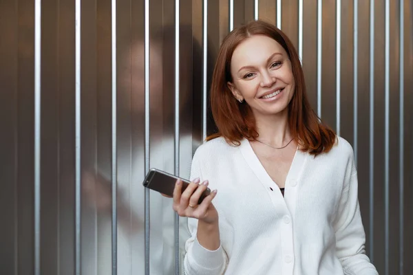 Удовлетворенная женщина-предприниматель с зубастой улыбкой, носит белый свитер, держит современный мобильный телефон, отправляет текстовые сообщения, ждет делового партнера, стоит в помещении с свободным пространством для вашего продвижения . — стоковое фото