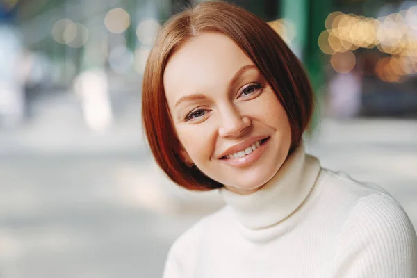 Image of attractive young woman with glad expression, looks directly at camera, has pleased look, wears turtleneck sweater, poses over blurred background, walks outside, expresses good emotions