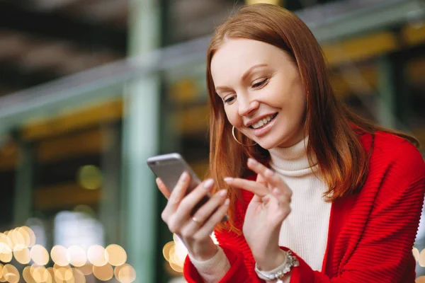 Положительно довольная женщина модель использует данные мобильного телефона, использует приложение чата, держит современный мобильный телефон, улыбается позитивно, одета в красное пальто, ждет ответа или звонка от бойфренда, имеет каштановые волосы — стоковое фото