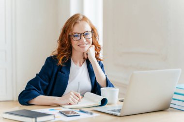 Başarılı ekonomist, gözlük, dizüstü bilgisayar modülleri denetler, Finans Web sitesinde bilgi okur, not defteri ve İçecek sıcak kahve yazıyor, işyerinde oturur. İş ve iş kavramı